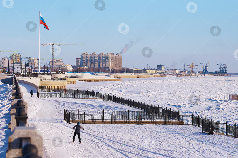 Скачать Набережная после снегопада. Зимний пейзаж в солнечный день. Флаг России и лыжник на набережной фотосток Ozero