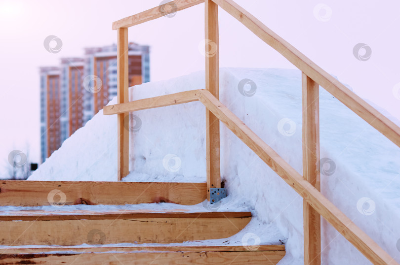 Скачать Ступени деревянной лестницы на ледяной горке. Зимнее сооружение из свежих досок, снега и льда. Раннее утро. Никаких людей. Жилые здания размыты на заднем плане фотосток Ozero