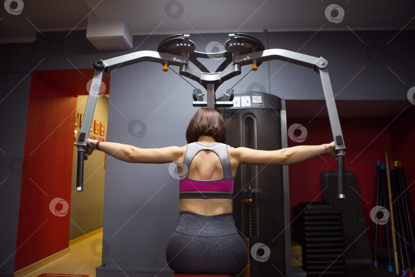 Скачать Женщина в спортивной одежде тренируется в тренажерном зале на тренажере для укрепления спины и рук. Спортивная форма, здоровый образ жизни, прокачка железа, поднятие тяжестей. Наращивание мышечной массы, потеря веса фотосток Ozero