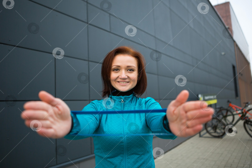 Скачать Женщина тренируется с фитнес-резинкой на городской улице. Укрепление кистей с помощью эспандера и утяжелителя. Фитнес, активный здоровый образ жизни, спорт на открытом воздухе фотосток Ozero
