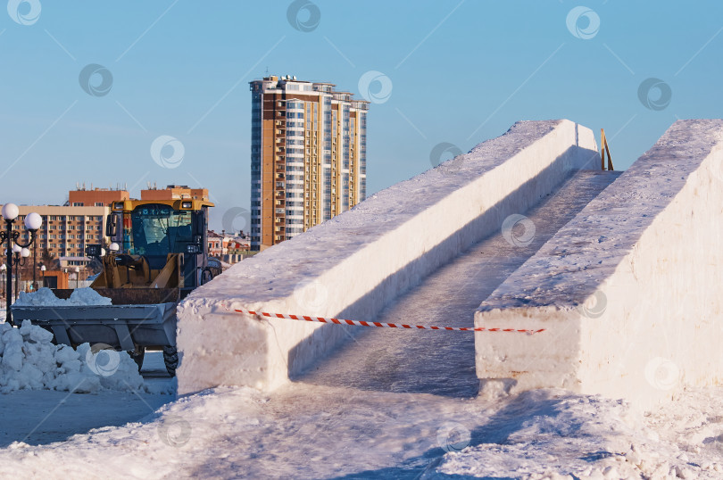 Скачать Снежная горка на городской площади. Трактор с ковшом расчищает детскую площадку. Стопорная лента закрывает слайд. Заливаем аттракцион льдом. фотосток Ozero