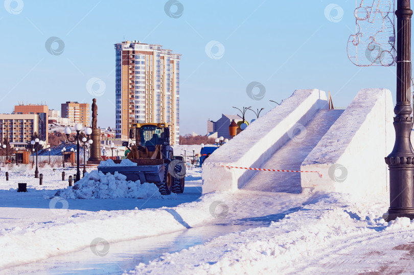 Скачать Снежная горка на городской площади. Трактор с ковшом расчищает детскую площадку. Заливаем аттракцион льдом. Подготовка к рождественским праздникам фотосток Ozero