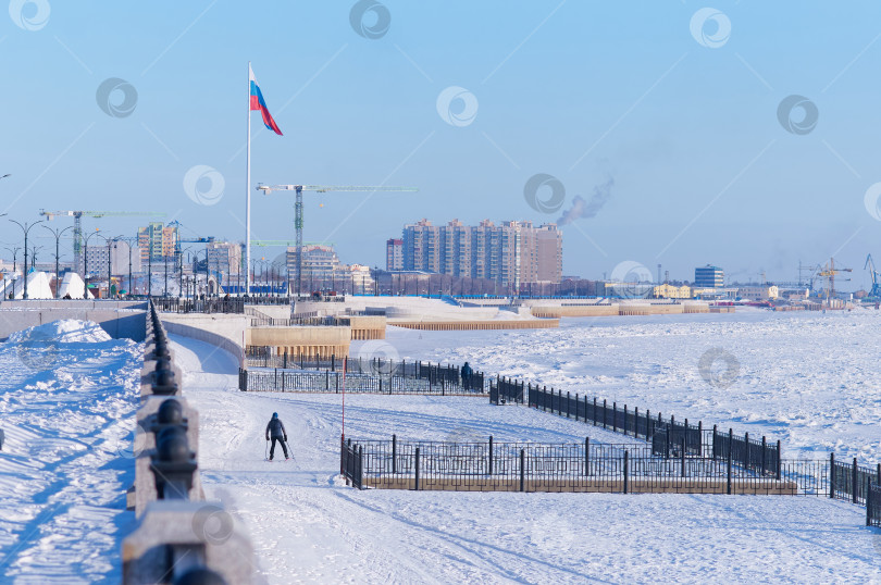 Скачать Snow-covered river embankment after a snowfall. Winter landscape on a sunny day. Flag of Russia and a skier on the promenade. Blagoveshchensk, Russia фотосток Ozero