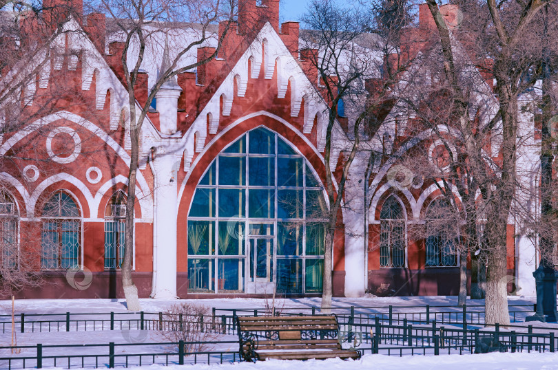 Скачать Facade of an old red brick building. Arched windows and white columns. Texture of frozen plants. Winter architectural landscape. Wooden bench and trees. фотосток Ozero