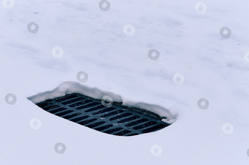 Скачать Black storm sewer grating on white snow in a snowfall. Winter symbol of urban infrastructure. фотосток Ozero