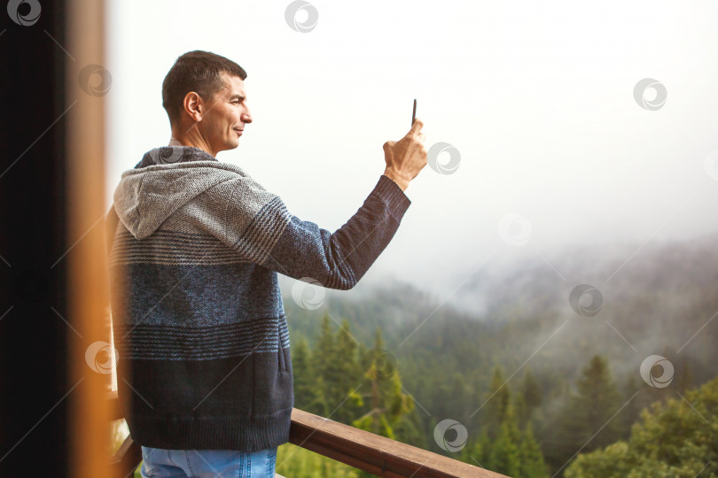 Скачать Молодой человек фотографирует на свой телефон прекрасный вид с балкона отеля, выходящего на горы. Вид на горы из окна, на ели, туман и холмы в доме. Селфи. Пространство для копирования фотосток Ozero