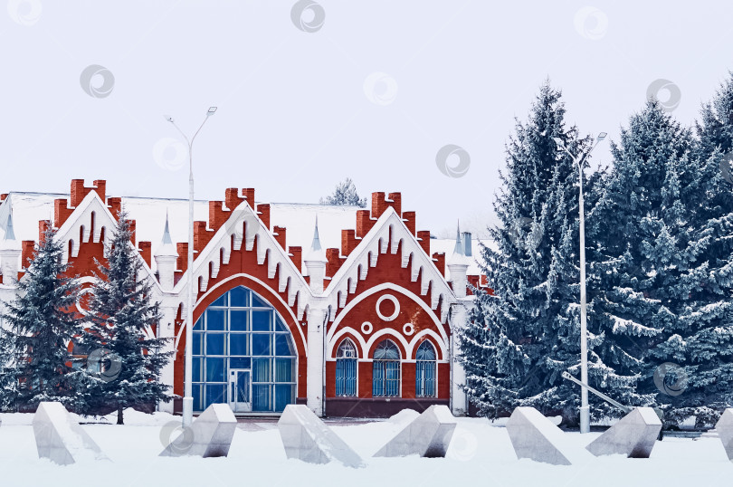 Скачать Winter architectural landscape. Fir trees with snow on the branches against the background of the facade of an old building made of red brick in the Gothic style. Red-white facade. фотосток Ozero