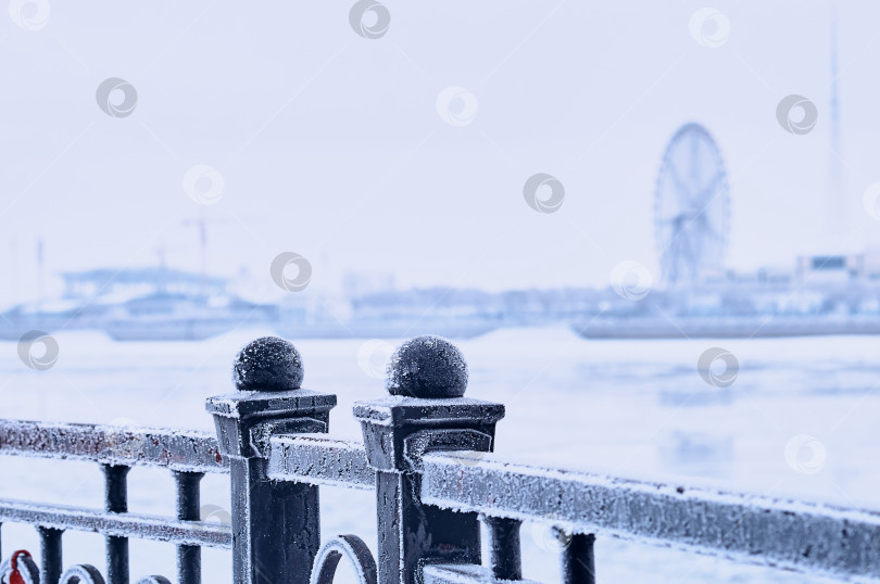 Скачать White frost on the black metal railings of the Amur river embankment on a winter morning. Ferris wheel silhouette in blur in the background. selective focus фотосток Ozero