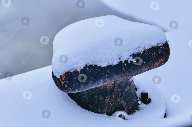 Скачать Mooring black cast-iron bollard under the snow on the river pier. Place of fastening of the mooring rope of the ship in the parking lot. фотосток Ozero