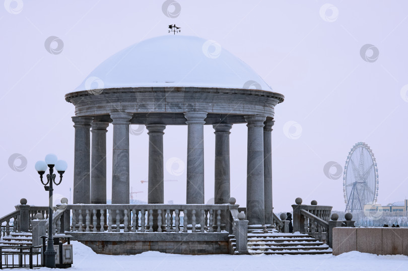 Скачать Granite gazebo in winter with columns and a balustrade against the backdrop of a Ferris wheel. Snow on the dome of the rotunda. фотосток Ozero