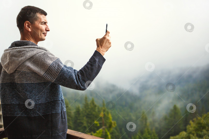 Скачать Молодой человек фотографирует на свой телефон прекрасный вид с балкона отеля, выходящего на горы. Вид на горы из окна на ели, туман и холмы в доме. Селфи. Пространство для копирования фотосток Ozero