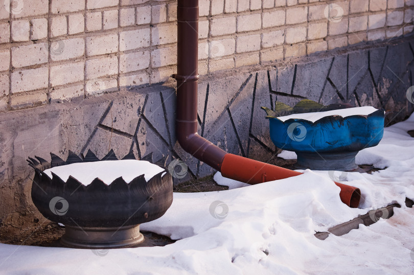Скачать Downpipe in winter on a brick wall with two homemade flower beds and snowdrifts. The concept of reusing car tires фотосток Ozero