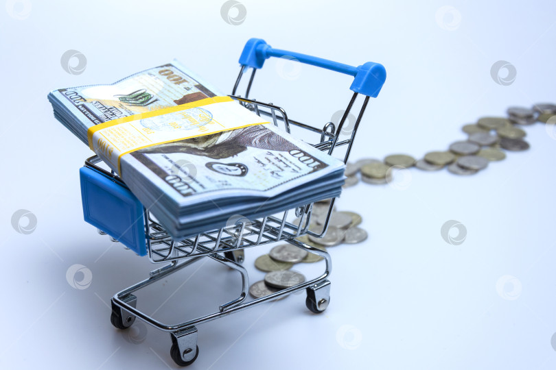 Скачать A supermarket trolley with a stack of hundred-dollar bills and scattered coins on a white background. фотосток Ozero