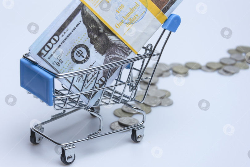 Скачать A supermarket trolley with a stack of hundred-dollar bills and scattered coins on a white background. фотосток Ozero