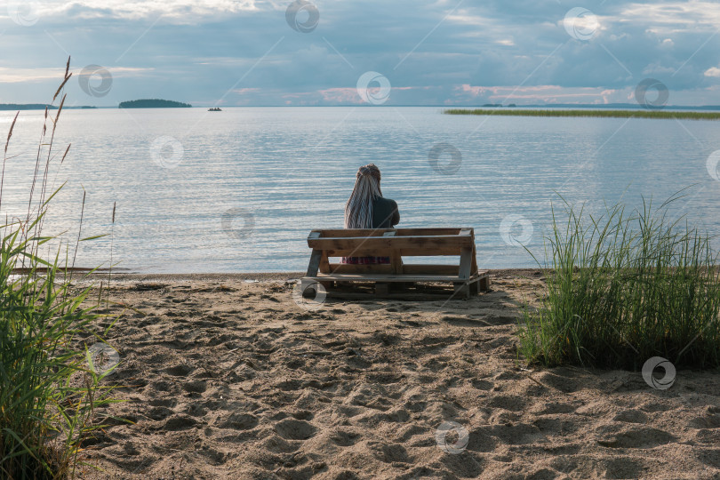 Скачать woman traveler with de braids hairstyle sitting on a bench made of old pallets on a sandy shore of a vast lake фотосток Ozero