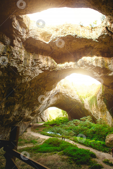 Скачать Пещера Деветашка в Болгарии - природная достопримечательность. Высокие своды огромной каменной пещеры с круглыми отверстиями наверху, туристическая дорога с забором внутри пещеры. фотосток Ozero
