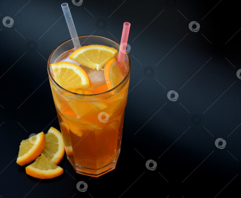 Скачать A tall glass of orange juice with ice and straws on a black background, next to pieces of ripe citrus. фотосток Ozero