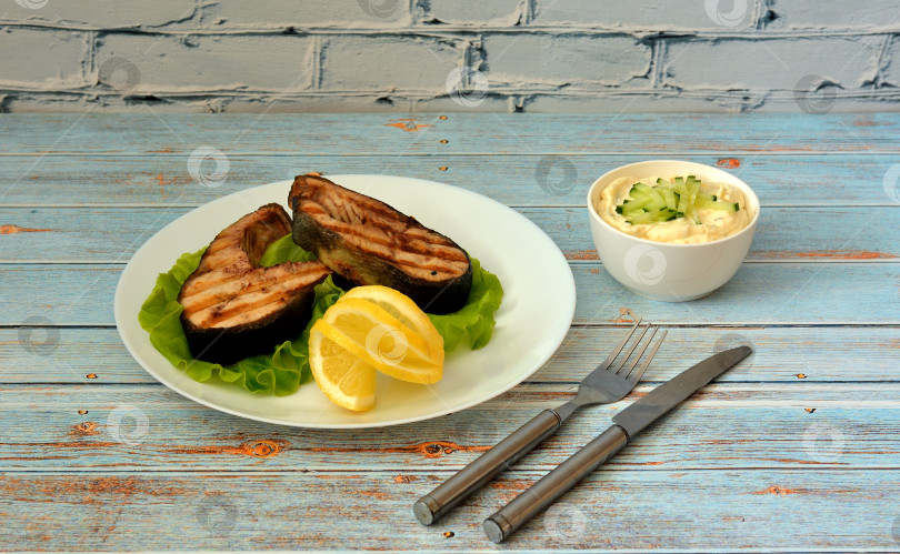 Скачать A plate with two salmon steaks on a lettuce leaf with lemon, sauce and cutlery on a wooden table. фотосток Ozero