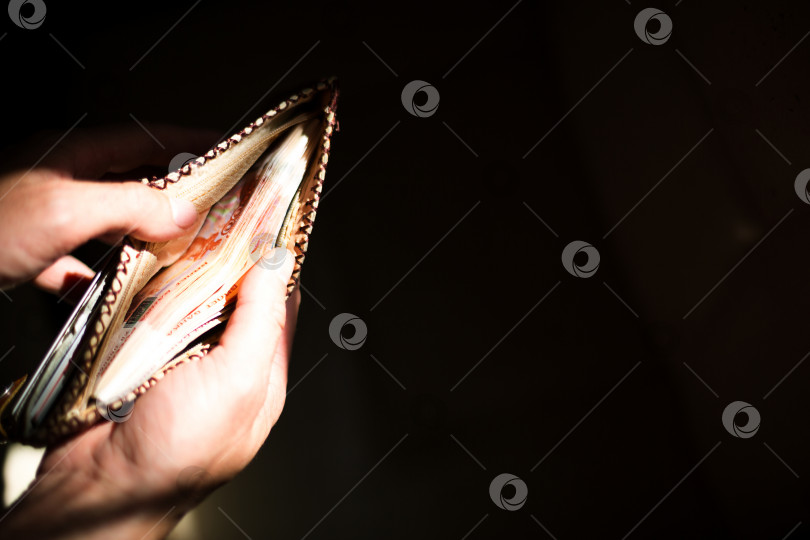 Скачать Кожаный бумажник, набитый российскими деньгами - 5000 купюр, пять тысяч рублей. Мужская рука держит открытый, переполненный бумажник на черном фоне в луче света. Пространство для копирования фотосток Ozero