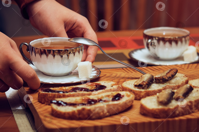 Скачать The girl puts two cups of hot tea and sandwiches from the tray on the table. Croutons with jam and sardines. Soft evening light, close-up. Cozy evening atmosphere. фотосток Ozero