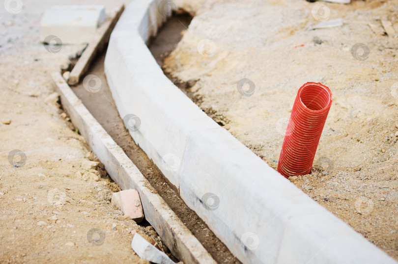 Скачать A red corrugated pipe for laying an electrical cable sticks out of the ground at a construction site. Curb stones are installed in a row using formwork and liquid concrete. фотосток Ozero