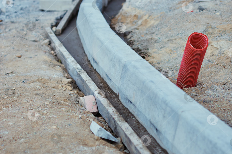 Скачать A red corrugated pipe for laying an electrical cable sticks out of the ground at a construction site. Curb stones are installed in a row using formwork and liquid concrete. фотосток Ozero