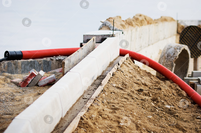 Скачать A red corrugated pipe for laying an electrical cable sticks out of the ground at a construction site. Curb stones are installed in a row using formwork and liquid concrete. фотосток Ozero