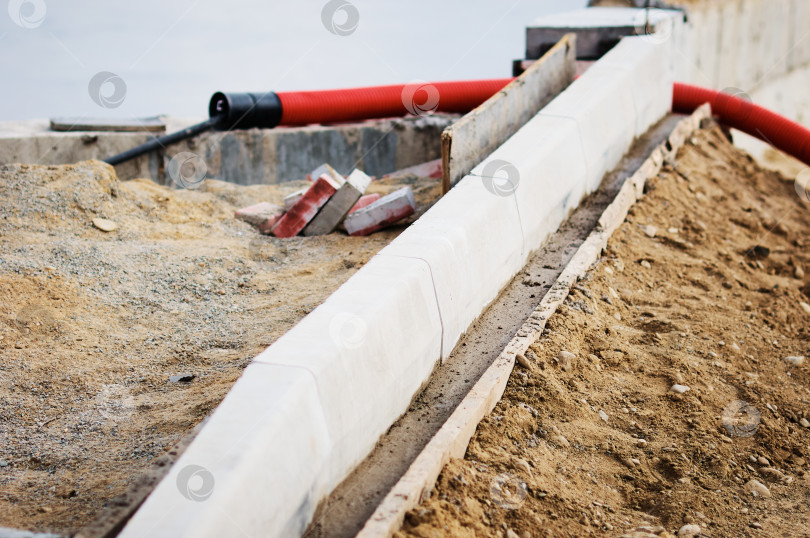 Скачать A red corrugated pipe for laying an electrical cable sticks out of the ground at a construction site. Curb stones are installed in a row using formwork and liquid concrete. фотосток Ozero