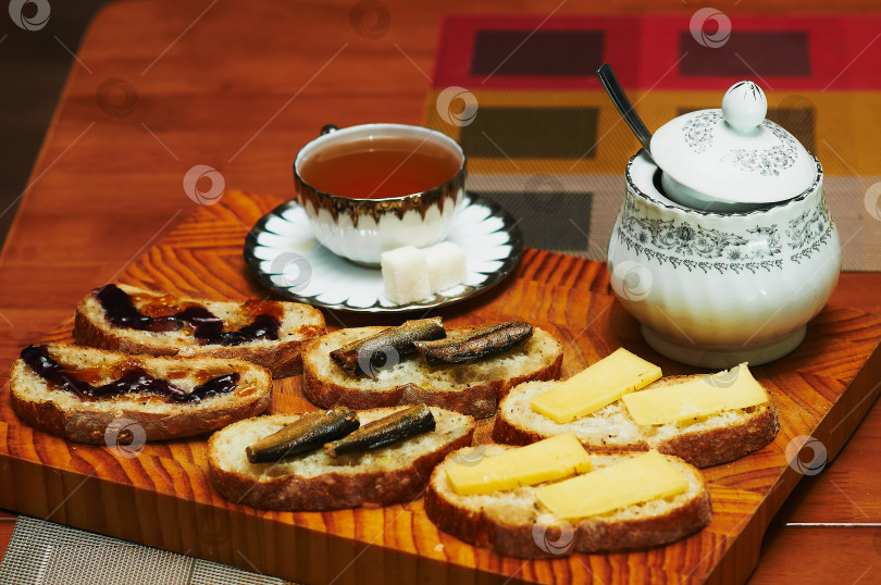 Скачать A cup of tea, a sugar bowl and sandwiches on a cutting board made of natural hevea wood. Croutons with jam, cheese and sardines. Soft evening light, close-up. Cozy evening atmosphere. фотосток Ozero