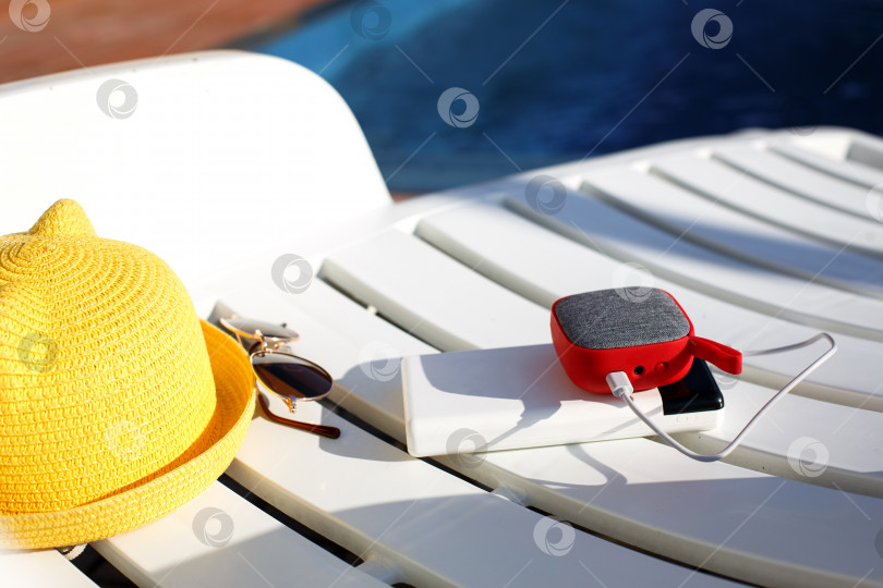 Скачать Музыкальный портативный динамик заряжается от блока питания через usb на шезлонге у бассейна с пляжными принадлежностями. Всегда на связи, гаджеты для путешествий, внешний аккумулятор для смартфона. Место для текста. Плоский лист фотосток Ozero