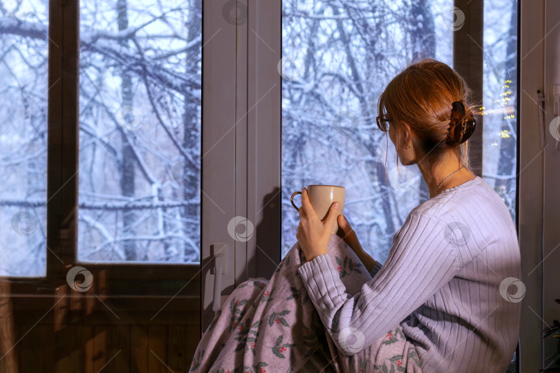 Скачать young woman sits on windowsill drinking from cup enjoying winter landscape and thinking. No face. Copy space фотосток Ozero