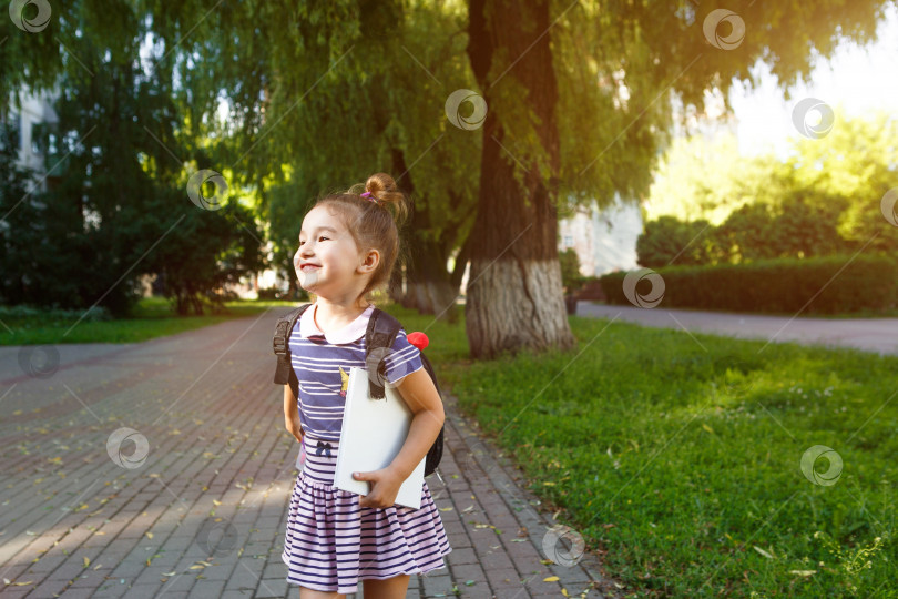 Скачать Маленькая девочка кавказской внешности в школьной форме с рюкзаком и книгой. Концепция возвращения в школу. Начальная школа, развивающие занятия для дошкольников. Пространство для текста фотосток Ozero