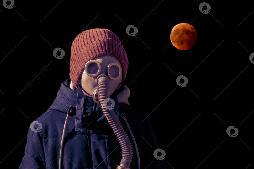 Скачать Парень в противогазе на фоне красной луны на черном небе во время лунного затмения. Теплая куртка и вязаная оранжевая шапочка. Концепция апокалипсиса фотосток Ozero
