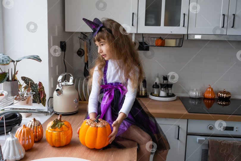 Скачать Ребенок украшает кухню дома к Хэллоуину. Девочка в костюме ведьмы играет с декором для праздника - летучими мышами, фонарем Джека, тыквами. Осенний уют в доме, кухня в скандинавском стиле, лофт фотосток Ozero