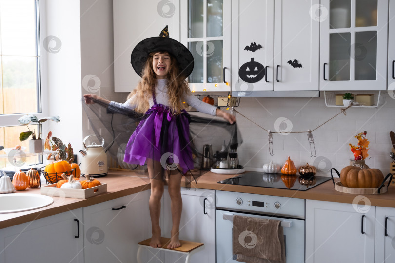 Скачать Ребенок украшает кухню дома к Хэллоуину. Девочка в костюме ведьмы играет с декором для праздника - летучими мышами, фонарем Джека, тыквами. Осенний уют в доме, кухня в скандинавском стиле, лофт фотосток Ozero