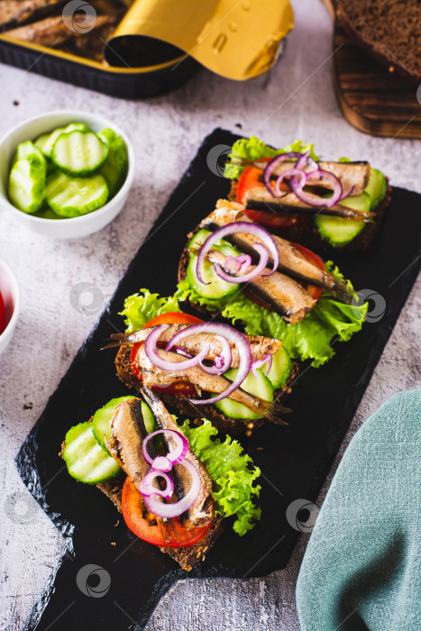 Скачать Бутерброды на ржаном хлебе с копчеными шпротами, овощами и зеленью на грифельной доске вертикальный вид фотосток Ozero