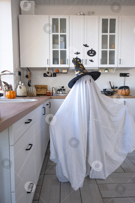 Скачать Ребенок в простынях с вырезом для глаз, похожий на костюм привидения, на кухне, украшенной к празднику Хэллоуин. Доброе маленькое забавное привидение. Вечеринка в честь Хэллоуина фотосток Ozero