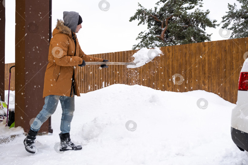 Скачать Мужчина зимой убирает снег лопатой во дворе дома на автостоянке. Снегопад, сложные погодные условия, машина буксует, выкапывая проезд фотосток Ozero