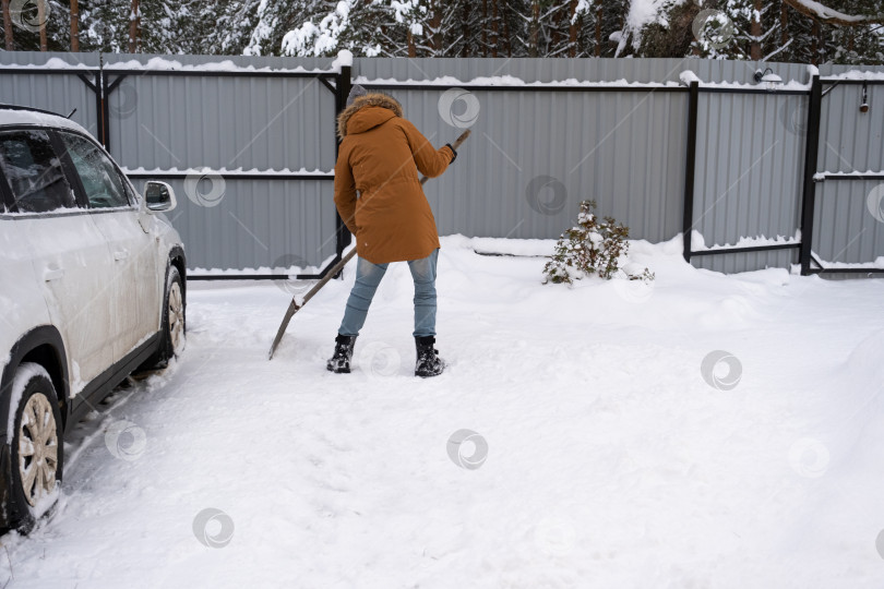 Скачать Мужчина зимой убирает снег лопатой во дворе дома на автостоянке. Снегопад, сложные погодные условия, машина буксует, выкапывая проход фотосток Ozero