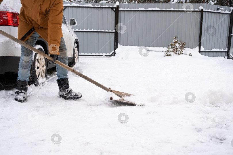 Скачать Мужчина зимой убирает снег лопатой во дворе дома на автостоянке. Снегопад, сложные погодные условия, машина буксует, выкапывая проход фотосток Ozero