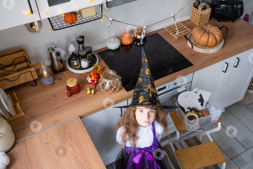 Скачать Ребенок украшает кухню дома к Хэллоуину. Девочка в костюме ведьмы играет с декором для праздника - летучими мышами, фонарем Джека, тыквами. Осенний уют в доме, кухня в скандинавском стиле, лофт фотосток Ozero