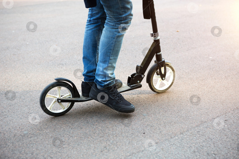 Скачать Ноги мужчины в джинсах и кроссовках на скутере в парке на асфальтированной дорожке. Спортивная прогулка, здоровый активный образ жизни, экологичный транспорт, дорожное движение фотосток Ozero