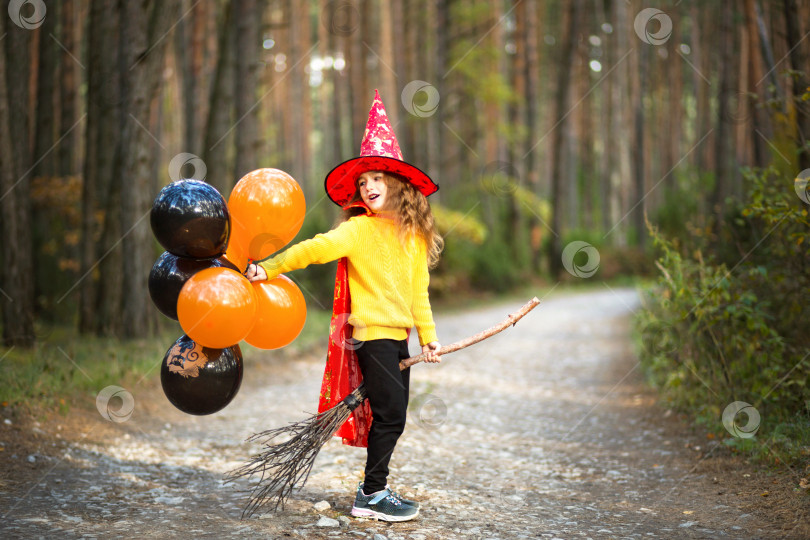 Скачать Девочка в костюме ведьмы и шляпе на метле с оранжевыми и черными воздушными шарами играет в осеннем лесу, собираясь на вечеринку в честь Хэллоуина фотосток Ozero