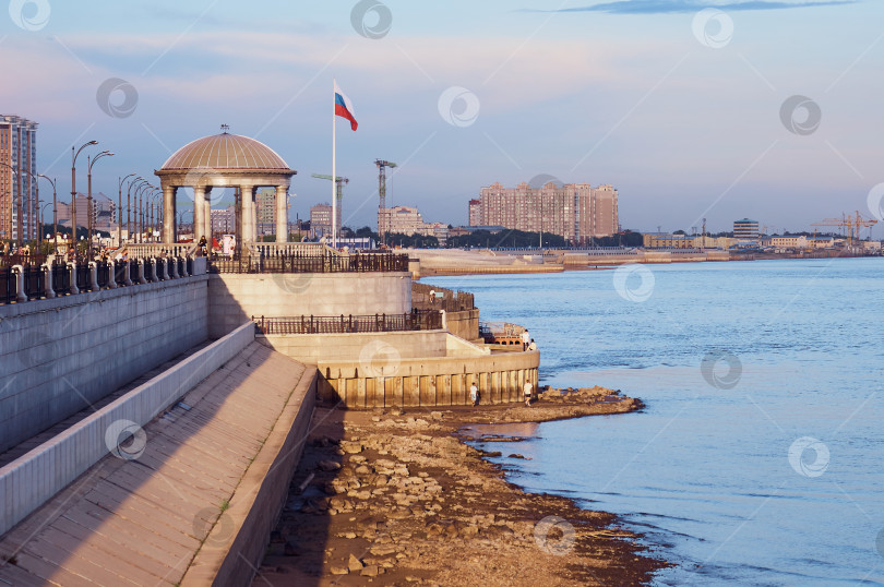 Скачать The embankment of the city with the rotunda and the Russian flag in the rays of the evening sun. The rocky shore of the Amur border river. фотосток Ozero