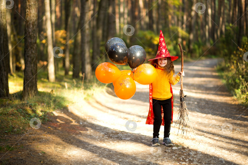 Скачать Девочка в костюме ведьмы и шляпе на метле с оранжевыми и черными воздушными шарами играет в осеннем лесу, собираясь на вечеринку в честь Хэллоуина фотосток Ozero