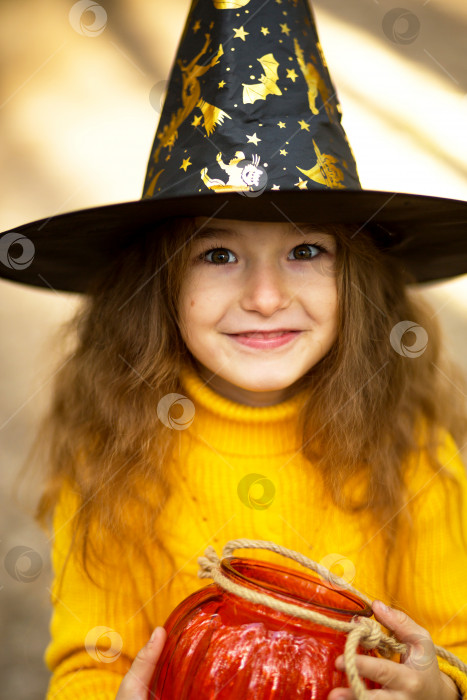Скачать Девочка в шляпе ведьмы, в желтом свитере и с тыквенным фонарем и ведерком для сладостей в осеннем лесу. Праздник Хэллоуина. Портрет, крупный план фотосток Ozero
