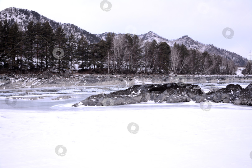 Скачать Каменные образования посреди замерзшего русла прекрасной реки, текущей по заснеженной горной долине пасмурным зимним утром. фотосток Ozero