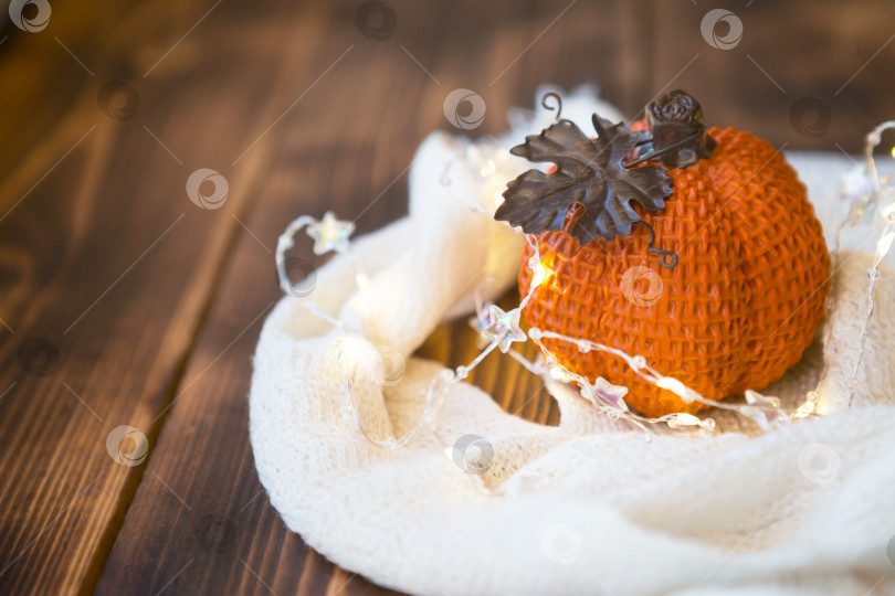Скачать Оранжевая тыква, завернутая в теплый белый плед, с гирляндами на деревянном столе в copyspace. Осеннее настроение, Хэллоуин, сбор урожая, день благодарения, домашний декор и уют. Избирательный фокус фотосток Ozero