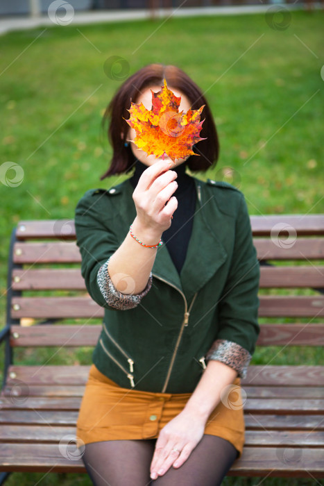 Скачать Женщина с желтым опавшим кленовым листом, закрывающим ее лицо. Осенний портрет без лица на скамейке в парке. Осеннее настроение фотосток Ozero