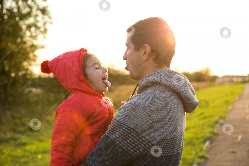 Скачать Маленькая девочка в красной куртке с капюшоном обнимает и высовывает язычок своего папу, улыбается. Счастливая семья, эмоции детей, день отца, яркие лучи солнца, кавказская внешность. Место для текста. фотосток Ozero